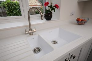 Corian sinks in Designer White stainless steel hot and cold faucet, window behind looking into the garden. On the window sill is a planted pot with two red flowers (not roses)